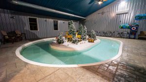 Indoor pool with a snowy Christmas scene on a small island featuring frosted trees and deer statues around a wooden chest in the center.
