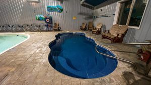 Indoor pool area with a curved blue pool, stone-tiled deck, and wooden lounge chairs along corrugated metal walls.