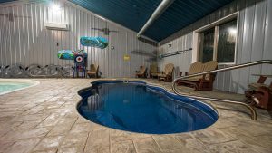 Indoor pool area with a curved blue pool, beige tiled deck, and brown Adirondack chairs along metal-paneled walls.