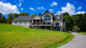 Large beige multi-gable house on a grassy, sloped lawn with a wooden deck and surrounding trees under a bright blue sky by a curved driveway.