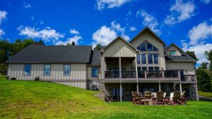 Two-story beige house with a large wooden deck and multiple chairs on a green lawn under a bright blue sky with scattered clouds.