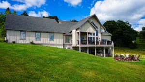 Exterior view of a beige, modern house on a grassy hill with a covered front porch and a wooden deck.