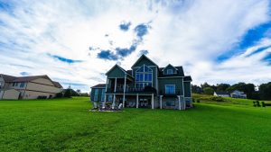 Green lawn in front of a modern green house with a large deck and outdoor seating around a fire pit, under a bright, cloudy sky.