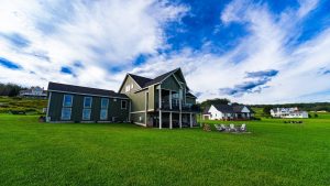 Green two‑story house with a wraparound balcony sits on a large grassy lawn under a bright blue sky with scattered white clouds.