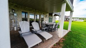 Back patio with a stone wall, several gray Adirondack chairs around a table under a covered porch, overlooking a green lawn.