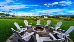Round stone fire pit surrounded by white Adirondack chairs on a green lawn, with houses and a blue sky in the distance.