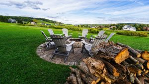 Circular array of gray Adirondack chairs around a stone fire pit on a brick patio, with stacked logs nearby and a green field beyond.