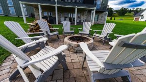 Circular arrangement of light-gray Adirondack chairs around a stone fire pit on a brick patio, with a wood pile nearby and a house with a covered patio in the background