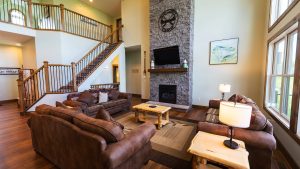 Cozy living room with brown leather sofas around a rustic wooden coffee table, stone fireplace, and wall of windows to the right.
