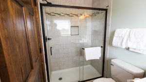 Glass-enclosed shower in a tiled bathroom with a white towel on the rack and a wooden door on the left side.