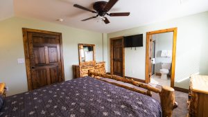 Rustic bedroom with log furniture, including a carved bed frame and dresser, two wooden doors, a wall-mounted TV, and a view into a bathroom.