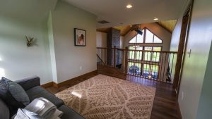 Living area with green walls, hardwood floor, and a staircase railing overlooking a bright vaulted-ceiling room with large windows and stone chimney.