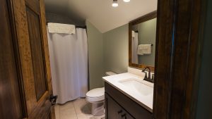 Bathroom scene showing a dark wood vanity with white countertop and dual-handle faucet, large framed mirror, and toilet beside a shower with a white curtain on the left.