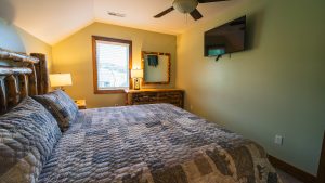 Rustic bedroom with a log-frame bed, gray quilt, two lamps, window with blinds, and a dresser with a mirror.
