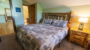 Rustic bedroom with a thick log bed, matching nightstand, and a warm lamp on the right; green walls and moose-pattern quilt.