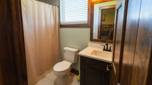Bathroom with toilet, sink, and dark wood vanity; white shower curtain along a corner and a window with blinds above the toilet.