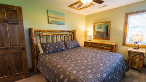 Rustic log bedroom with a large woody bed, matching dresser and mirror, green walls, and a window with blinds reflecting natural light.