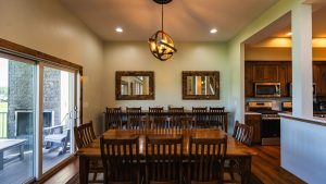 Dining room with a long wooden table and many chairs, two square mirrors on the wall, and a globe-style chandelier; sliding glass door to the patio on the left.