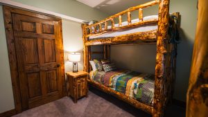 Rustic log bunk bed in a cozy cabin room with a quilted bedspread and a wooden nightstand with a lamp in the corner.