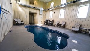 Indoor pool area inside a metal-paneled room, with several Adirondack chairs along the wall and tall windows letting in light.