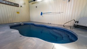 Indoor pool area with a curved blue pool and surrounding concrete deck; beige metal walls and a metal handrail on the right.