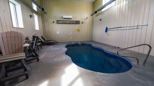 Indoor pool area with curved blue water and gray concrete floor, surrounded by wooden-paneled walls and lounge chairs along the left wall.