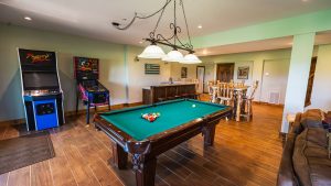 A cozy game room with a green pool table, hanging light fixture, and wooden bar surrounded by high chairs.
