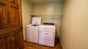 Laundry closet with a white top-load washer (lid open) and a matching front-load dryer side by side, under a metal wire shelf, next to a wooden door.