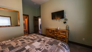 Bedroom with a wooden dresser under a wall-mounted flat-screen TV, cords hanging nearby and a vase on the dresser.
