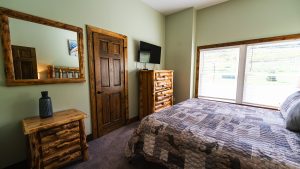 Rustic bedroom with log-style furniture, a patterned quilted bed, and a wooden dresser by a green wall beneath a windowed wall.