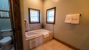 Bathroom scene with a white corner tub, two wooden-framed windows, and a towel rack with white towels; a sign on the left reads 'flush wash floss brush'.