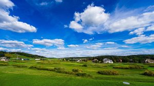 Wide view of a green suburban landscape with a row of houses along the horizon under a bright blue sky dotted with white clouds.