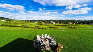 Circular fire pit surrounded by white Adirondack chairs on a green lawn, with a suburban golf course and houses in the distance beneath a bright blue sky.