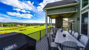 Outdoor deck with a long dining table and many gray chairs, grill covered in foreground, overlooking a green golf course and houses under a blue, cloudy sky.