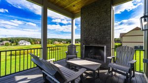 Covered deck with a stone fireplace, dark wood Adirondack chairs, and a wooden table, overlooking a green lawn and suburban houses under a bright blue sky.
