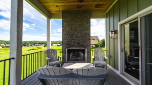 Covered patio with a tall stone fireplace centerpiece, four gray Adirondack chairs, and a black railing overlooking a green suburban lawn.