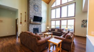 Spacious living room with a tall stone fireplace, mounted TV, and large windows that fill the space with light; brown leather couches surround a wooden coffee table.