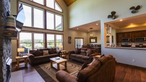 Open living room with large vaulted windows, a stone fireplace on the left, and brown leather sofas facing a wooden coffee table. The kitchen is visible to the right.