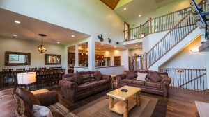 Open-concept living room with brown leather sofas, wooden coffee table, and a two-story staircase with iron balusters; dining area and kitchen in the background.