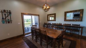 Rustic dining room with a long wooden table and many chairs; circular chandelier overhead, and sliding glass door to a deck outside with a green lawn view.