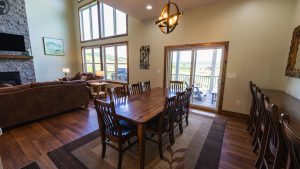 Spacious dining area with a long wooden table and multiple chairs, adjacent living space, large windows, and a stone fireplace on the left, overlooking outdoors.