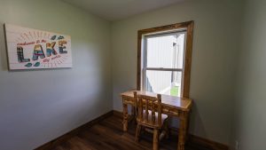 Wooden desk and chairs sit in a small green-walled room; a lake-themed sign reading 'Welcome to the Lake, stay awhile' hangs on the wall beneath a window with a wooden frame.