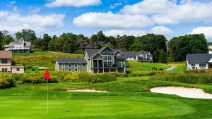 Sunny golf course scene with a red flag on a green, houses on a hill in the background and trees under a blue sky.