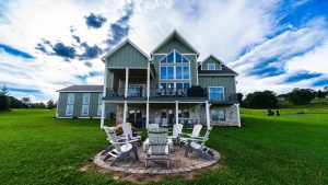 Green multi-story house with a large glass-front and a wraparound deck, set on a lush lawn with Adirondack chairs around a circular brick fire pit in the foreground.