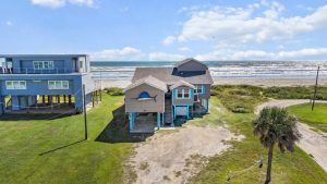 Aerial view of blue beach houses on stilts along the shoreline with the ocean in the background.