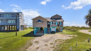 Beach houses on stilts at a coastal area with a sandy path and blue sky. A large fish sculpture sits on the central house.