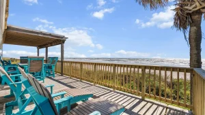 Beachfront wooden deck with bright turquoise Adirondack chairs overlooking the ocean under a blue sky with scattered clouds.