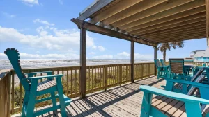 Beach deck with turquoise Adirondack chairs under a wooden pergola facing the ocean and blue sky with scattered clouds.
