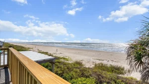 Beach scene with blue sky, white clouds, and calm ocean seen from a wooden railing on a sandy shore.