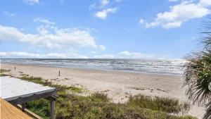 Wide view of a sunny beach with a calm sea, blue sky and scattered clouds, sandy shore bordered by dune grasses and a wooden railing in the foreground.